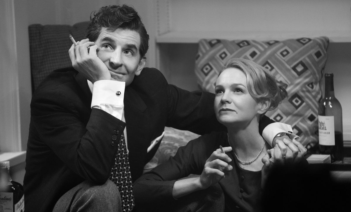 Bradley Cooper as Leonard Bernstein and Carey Mulligan as Felicia Montealegre sit together, smoking and laughing at someone off-screen, in a black-and-white image from Netflix's Maestro