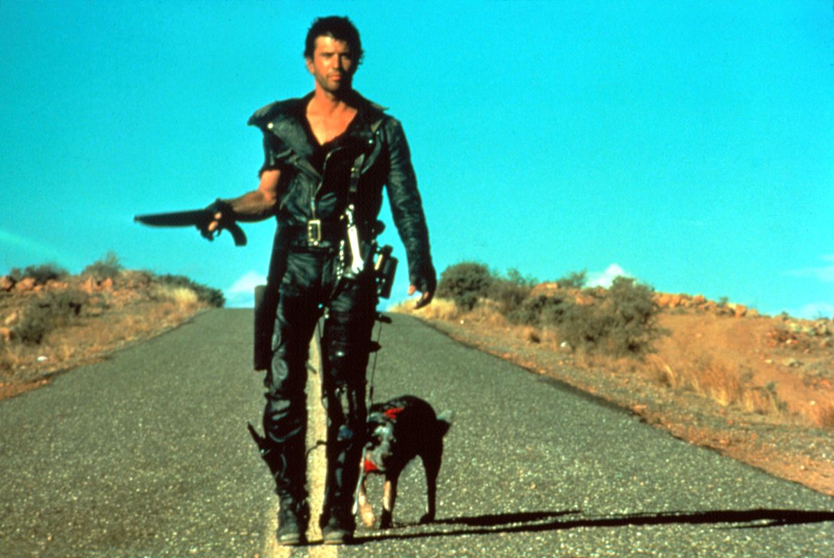 Mel Gibson as Mad Max, dressed all in black leather and carrying a short shotgun, walks down the road with a dog in Mad Max 2: The Road Warrior.