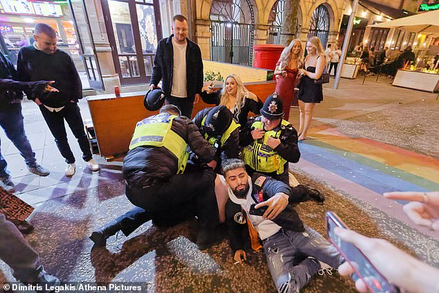 In this shot, one of the men now lies on the ground taking a selfie for the officers, while another holds a police hat and a third poses for the camera in front of a large group of onlookers