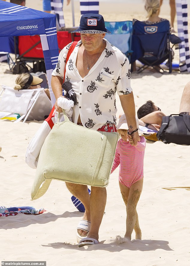 Karl wore a white Hawaiian shirt and red patterned board shorts, as well as a blue bucket hat as he took over from Jasmine and led their daughter to the spot with the rest of the family further down the beach.