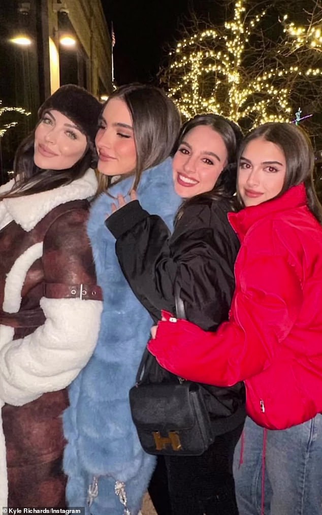 The four posed in front of trees illuminated by festive Christmas lights, with one wearing a blue fur coat, one in black, one in black and one in a brown leather variant with white Sherpa trim