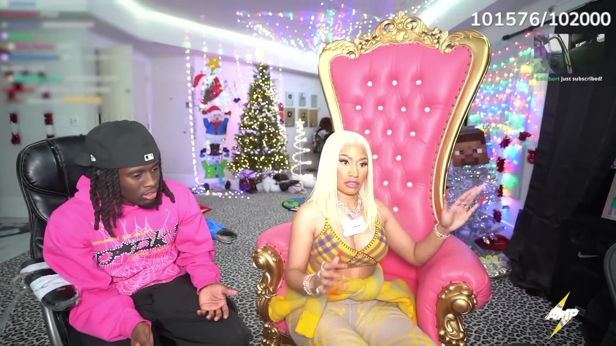 A still from a stream with Kai Cenat and Nicki Minaj.  She sits on a pink throne-like chair and Kai sits next to her in an office chair.  Minaj looks at the chat and Kai looks down as he talks to her.  They are in a basement with a Christmas tree in the background.