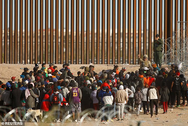 Migrants gather at the border wall after crossing the Rio Bravo River as a Border Patrol agent commands the group