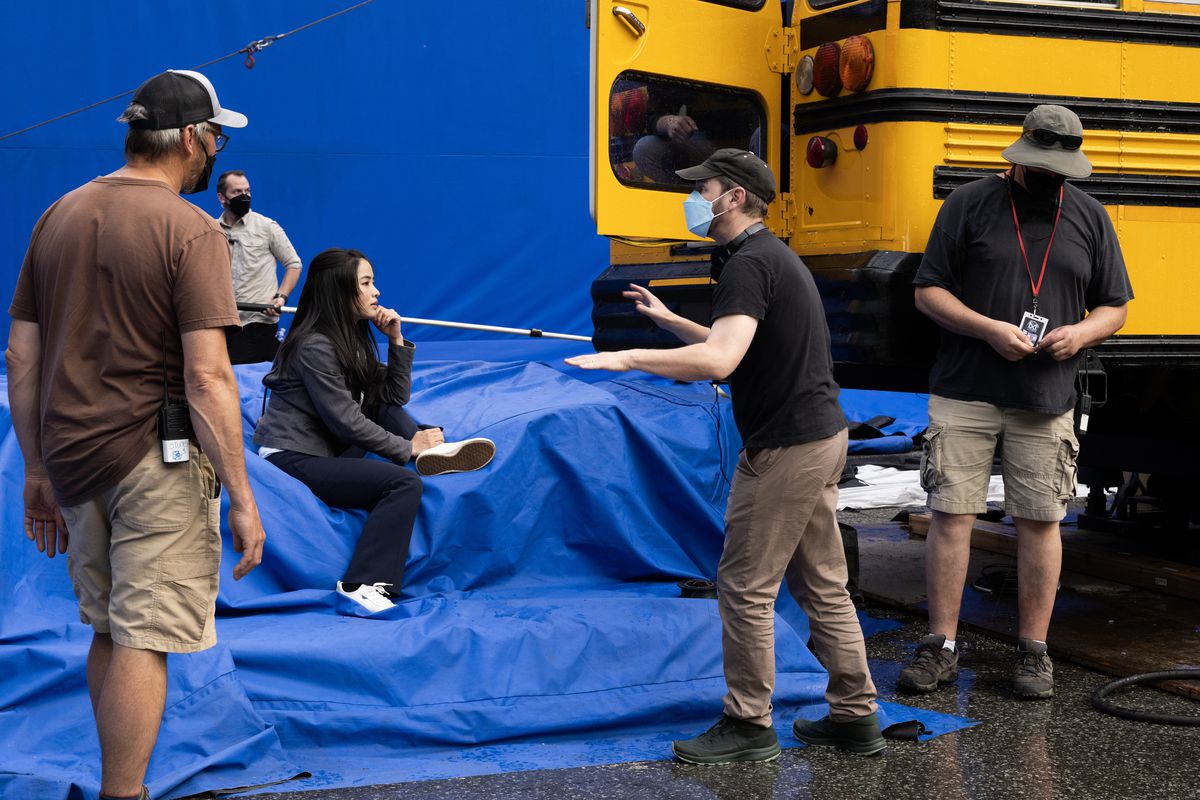 Matt Shakman talks to Anna Sawai on the set of Monarch;  she sits on a giant blue screen next to a yellow school bus