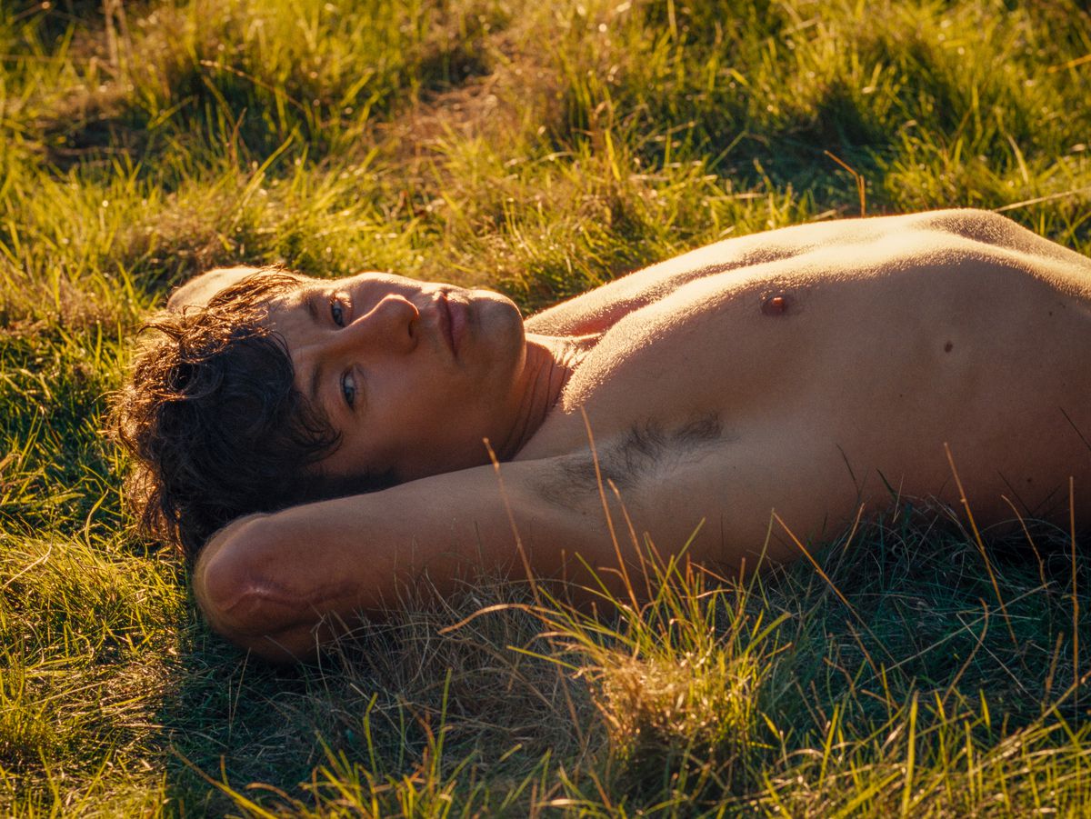 Oliver (Barry Keoghan), shirtless and lying on his back with an arm behind his head in a sunny meadow in Emerald Fennell's Saltburn