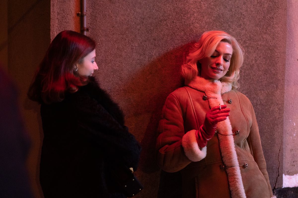 Anne Hathaway, in a blonde wig and sheepskin coat, smokes leaning against a neon-drenched wall as Rebecca looks on as Thomasin McKenzie looks on in the film Eileen.