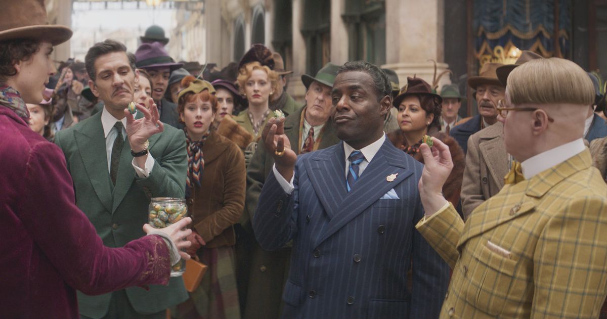 Mathew Baynton in a green suit, Paterson Joseph in a blue suit and Matt Lucas in a yellow suit talking to Willy Wonka