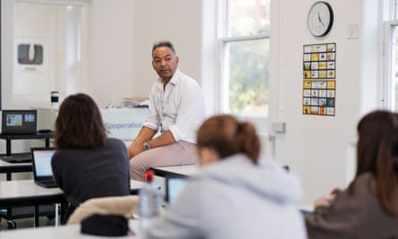 Steve Green is taking a biology class at GCSE – he sits in a classroom opposite three girls who we see from behind;  he wears an open white shirt and pale jeans, one of the girls wears a hoodie and a relaxed atmosphere is suggested