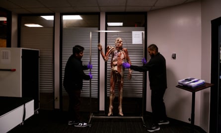 In a mostly dark, fluorescent-lit conference room, two young men on either side hold a metal frame containing a synthetic body that shows muscle and tissue.