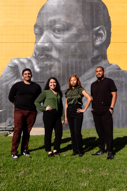 Four young Brown people stand upright in a line, in the sun so their shadows fall behind them, the women in the center with one arm on a hip, in front of a detailed grayscale mural of Martin Luther King Jr.  on a yellow background.