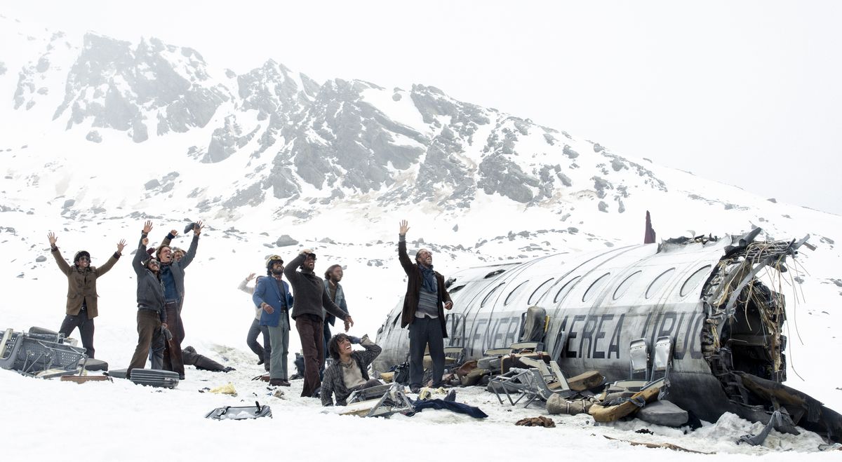 A group of survivors, seen from a great distance as tiny figures, stand in the snow next to their fragmented plane and wave to something off-screen in Netflix's Society of the Snow.