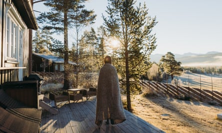 Woman in a blanket on a terrace next to a wooden building with sun shining through the pine trees and mountains in the distance in Norway's Rondane National Park 