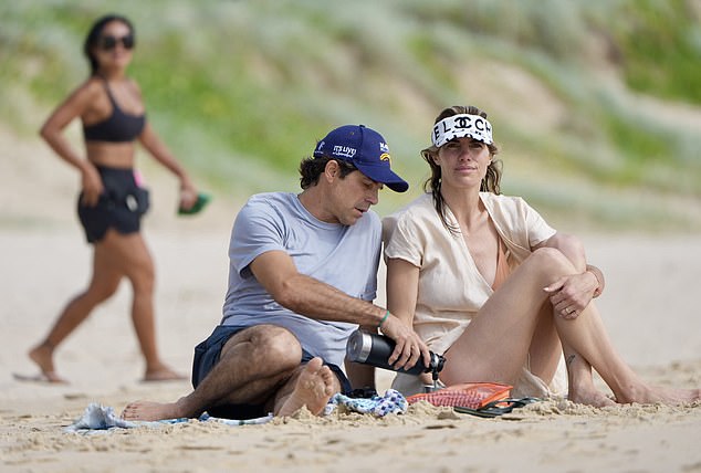 Delfina had arrived earlier at the beach wearing a loose cream-colored cover-up shirt and a black and white sun visor