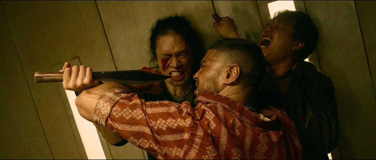 Nassim Lyes struggles with two opponents in the elevator fight scene of Mayhem!, specifically a bloodied man who tries to stab Nassim in the face.