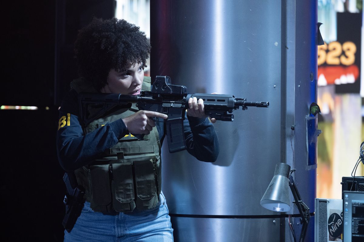 Emmy Raver-Lampman, wearing a bulletproof vest and FBI uniform, aims an assault rifle at The Beekeeper.