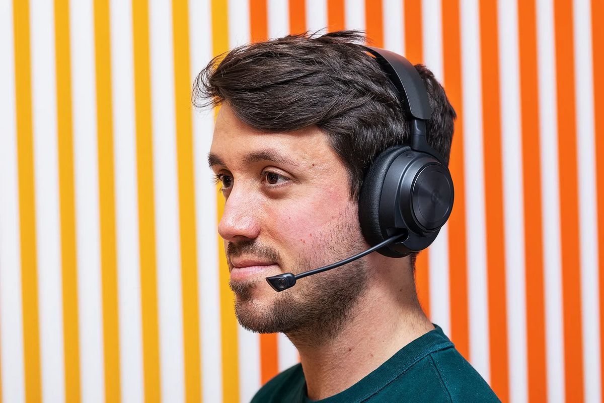 The SteelSeries Arctis Nova Pro Wireless is worn by the author, Cameron Faulkner.  He stands in front of a wall with a gradient pattern from warm yellow to orange.