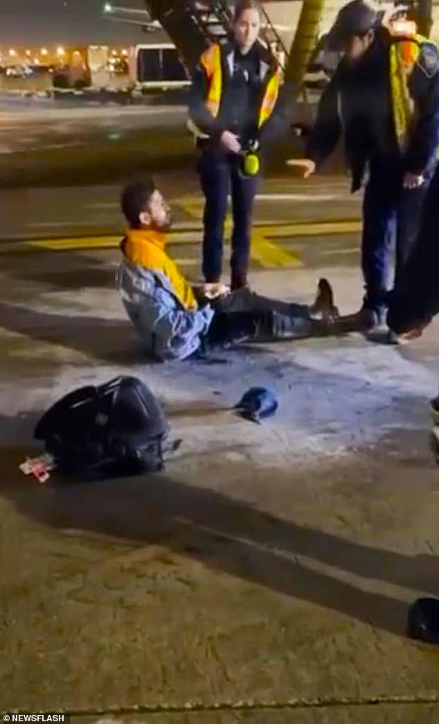 Airport staff, dressed in high-visibility clothing, appear to tell the man to lie still after he falls from the plane