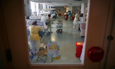 A view through a window at a row of incubators near a wall of windows, with mostly women visibly wearing yellow protective smocks and masks.