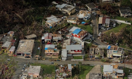 An aerial view of one-story wooden buildings along an asphalt road with fallen red roofs and blue walls.