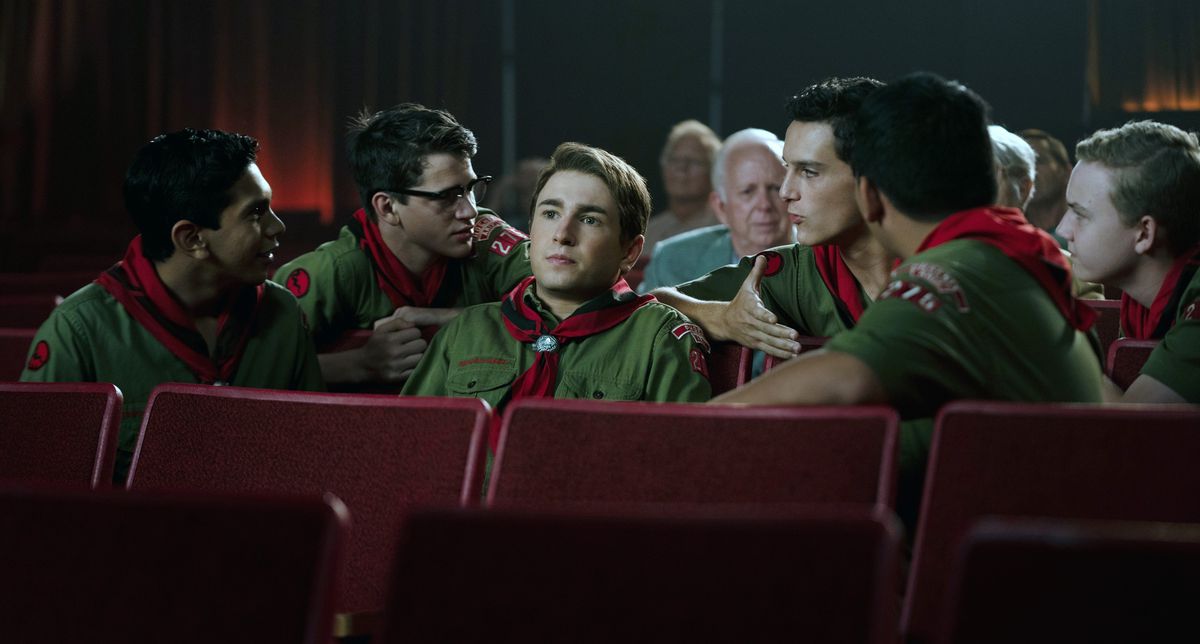 Sammy Fabelman (Gabriel LaBelle), a teenage boy in a green scout uniform with a red neckerchief, sits in a movie theater watching his latest film project while all his scout friends sit around him and talk to him in Steven Spielberg's The Fabelmans