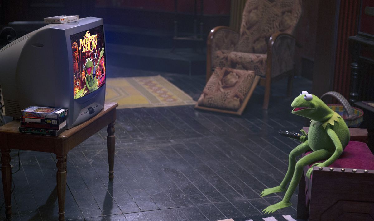 Kermit the Frog sits on a low, red upholstered couch and watches The Muppet Show on TV in Muppets Most Wanted