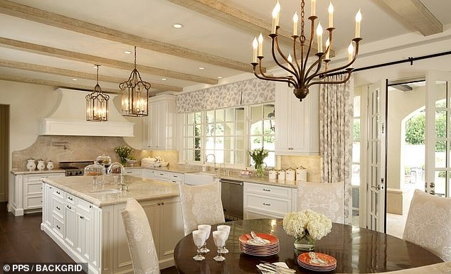 The kitchen is decorated with golden ceiling beams and features an island and beige marble worktops, with a matching backsplash