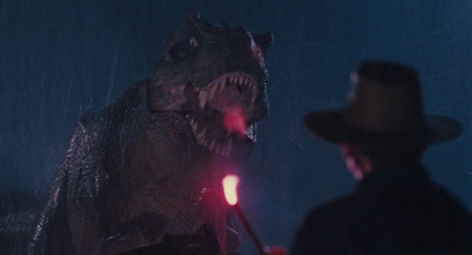 A man in a hat holds a flare in front of a roaring Tyrannosaurus Rex in Jurassic Park.