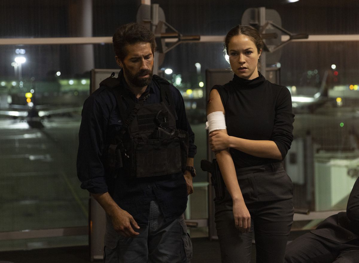 Scott Adkins stands next to an injured Hannah Arterton, with a bandage on her arm, at the airport in One More Shot.