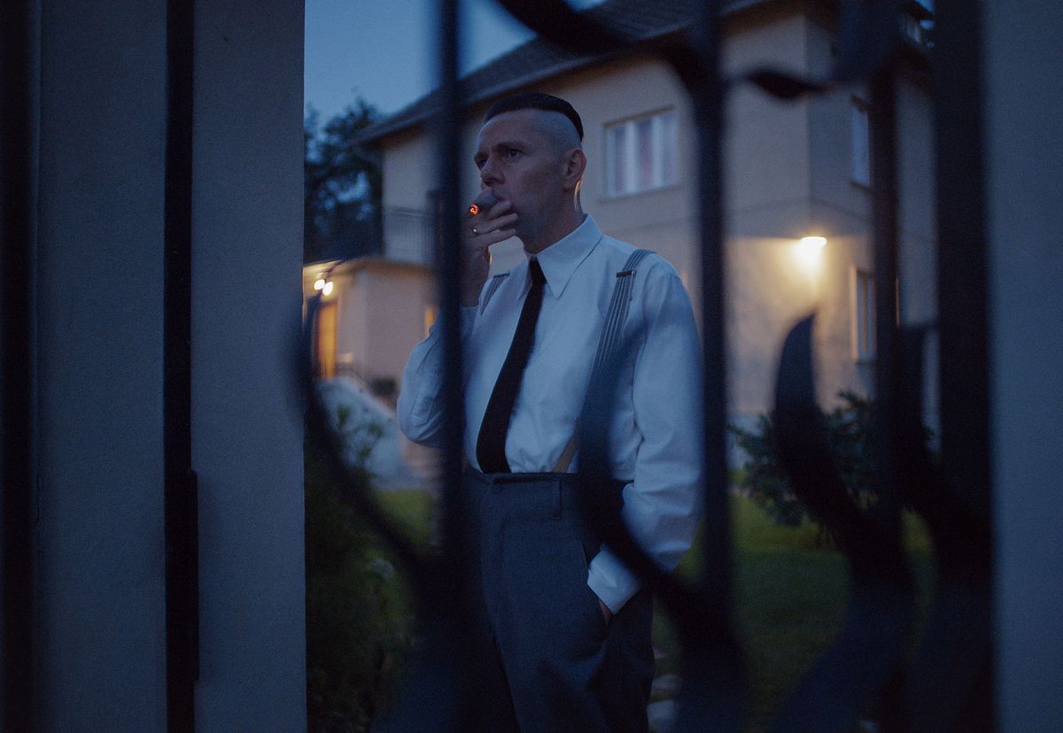 Christian Friedel as Rudolf Höss standing inside a fence smoking a cigarette in The Zone of Interest