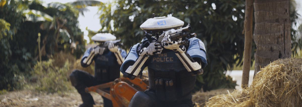 Two robot police officers, crouched in a tropical setting, aim shotguns in The Creator.