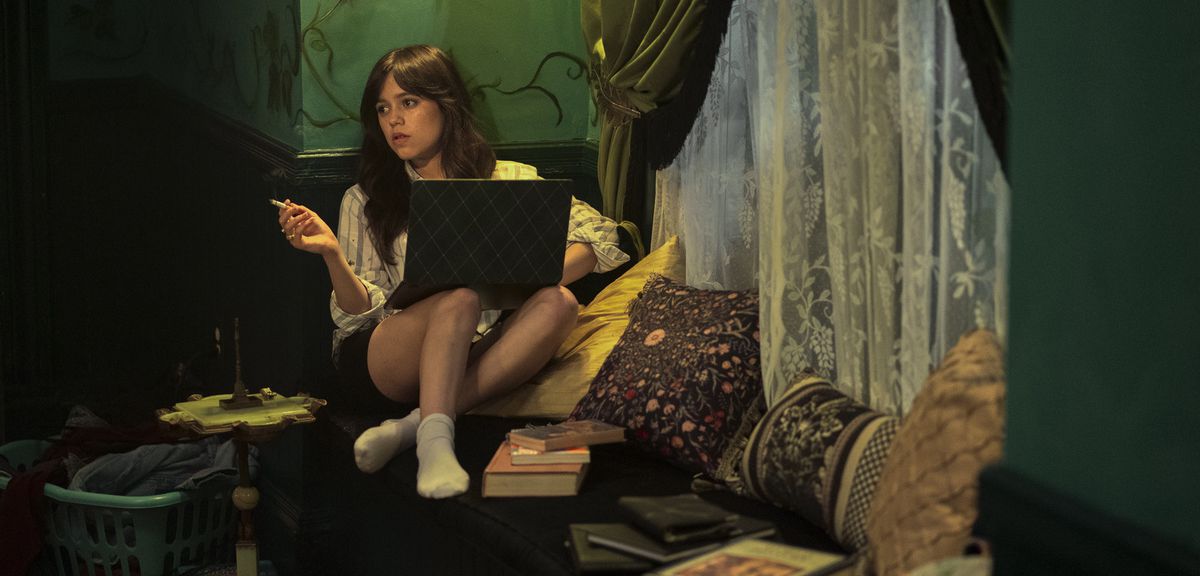 Cairo (Jenna Ortega) sits and smokes with a laptop on her lap and her legs folded in a chair, surrounded by pillows, books and a gauzy lace curtain in Miller's Girl