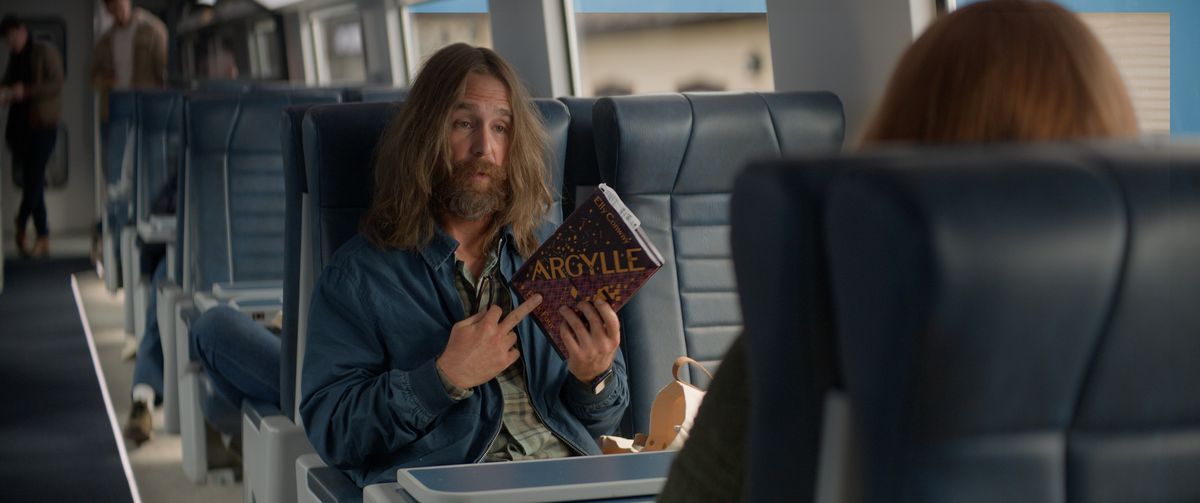 Sam Rockwell, with long hair and beard, holds a copy of the book Argylle by Elly Conway in the movie Argylle