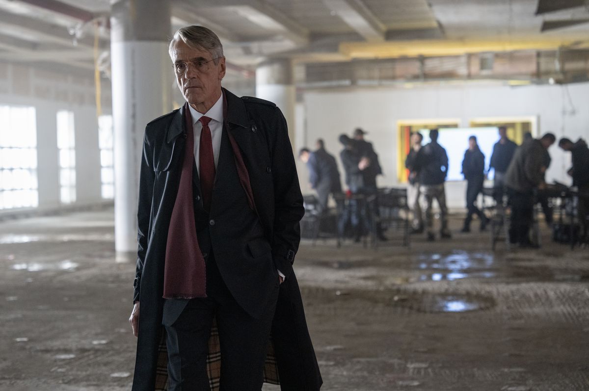 Jeremy Irons, wearing a long coat, walks toward the camera and away from the uniformed people in what appears to be a parking garage at The Beekeeper.