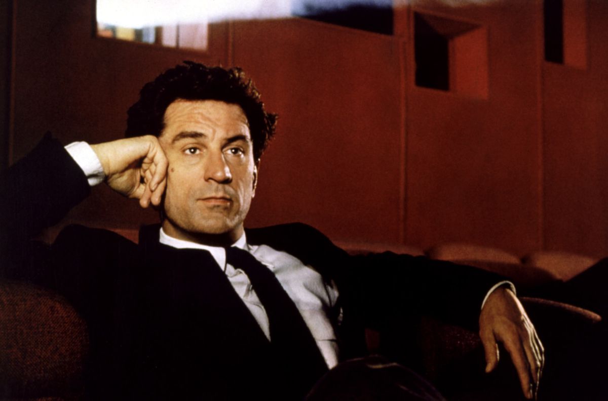 Hollywood director David Merrill (a young Robert De Niro, in a black suit and tie and a white-collared shirt) sits in a red-walled movie theater and watches a movie with the projector light behind him in 1991's Guilty by Suspicion
