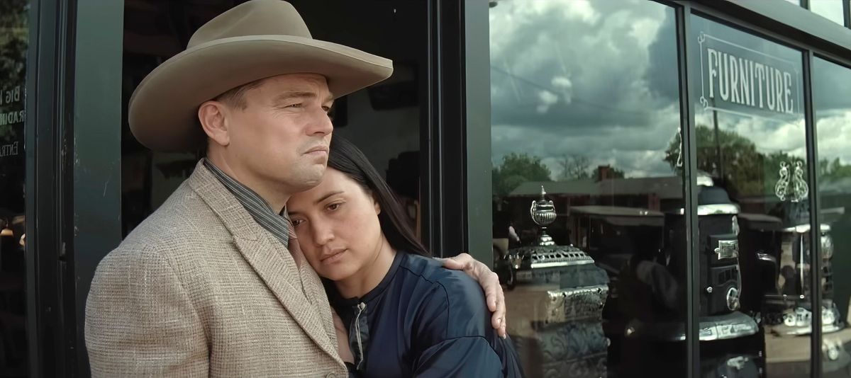 Ernest (Leonardo DiCaprio, in a brown suit and cowboy hat) comforts his wife Mollie (Lily Gladstone) as she leans against him in the window of a furniture store in Killers of the Flower Moon