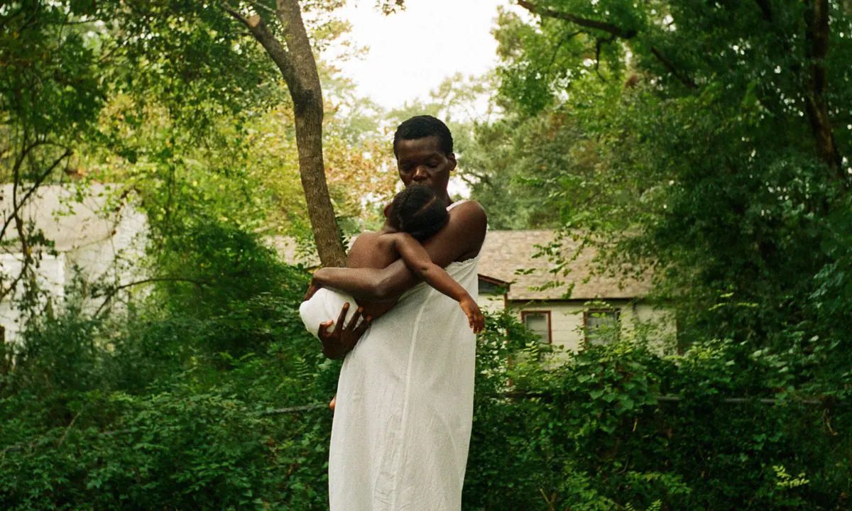 A woman (Sheila Atim) in a white dress holding a child in front of a thicket of trees and shrubs with a house in the background in All Dirt Roads Taste of Salt.