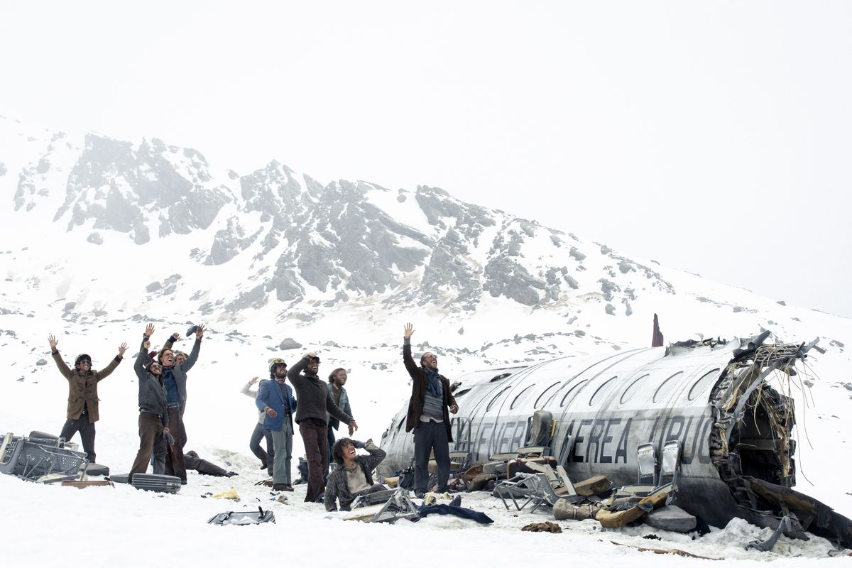 A group of men and women standing next to the wreckage of an airplane with a snowy mountain in the background in The Society of the Snow.