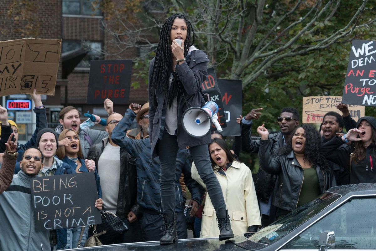 A young woman standing on the hood of a car in front of a group of protesters in The First Purge.