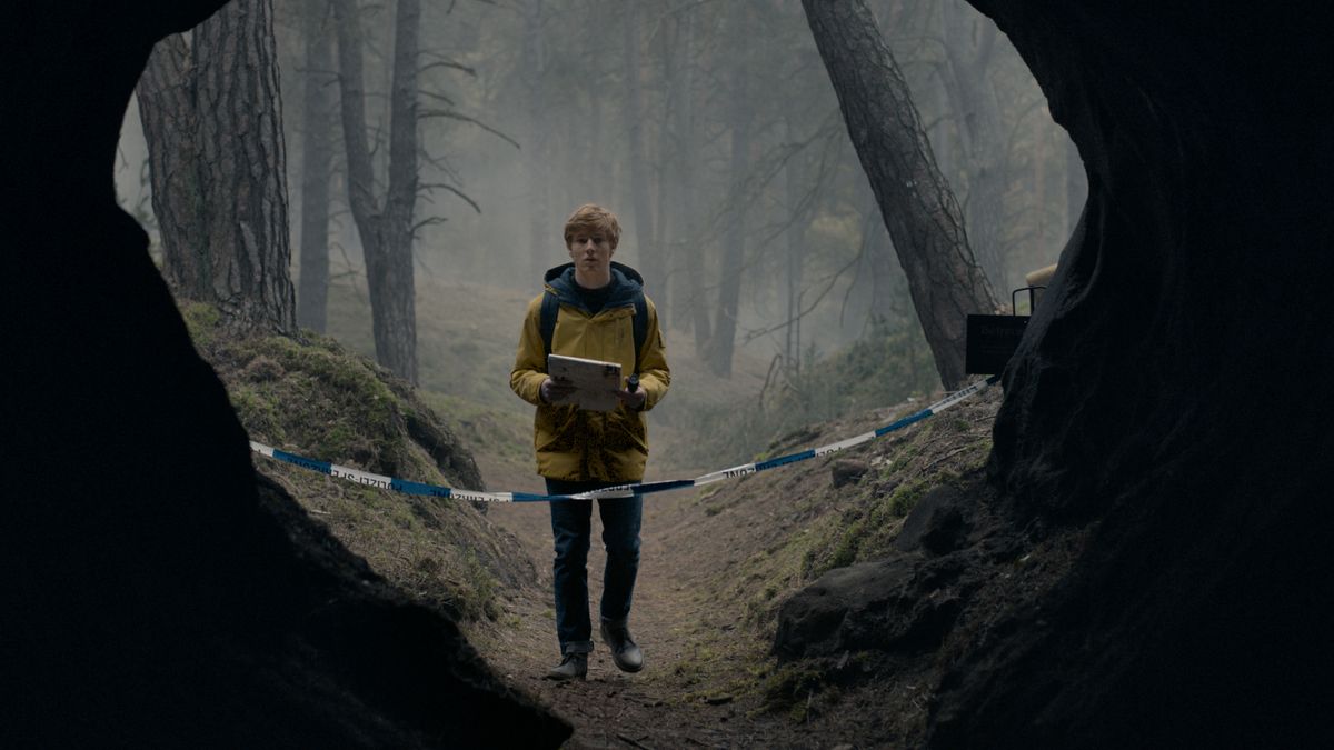 A young man, dressed in a yellow raincoat, stands in front of the separate entrance to a large cave in a forest in the dark.