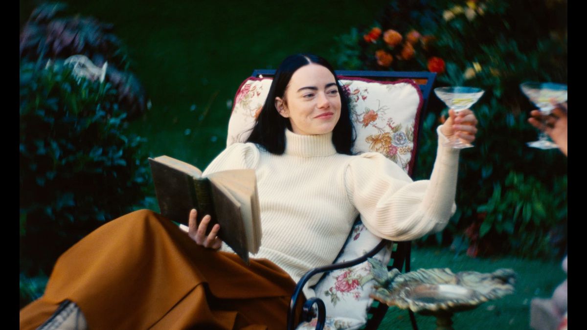 Emma Stone as Bella Baxter, lounging in a lawn chair, holding a book, smiling and raising a cocktail glass in Poor Things