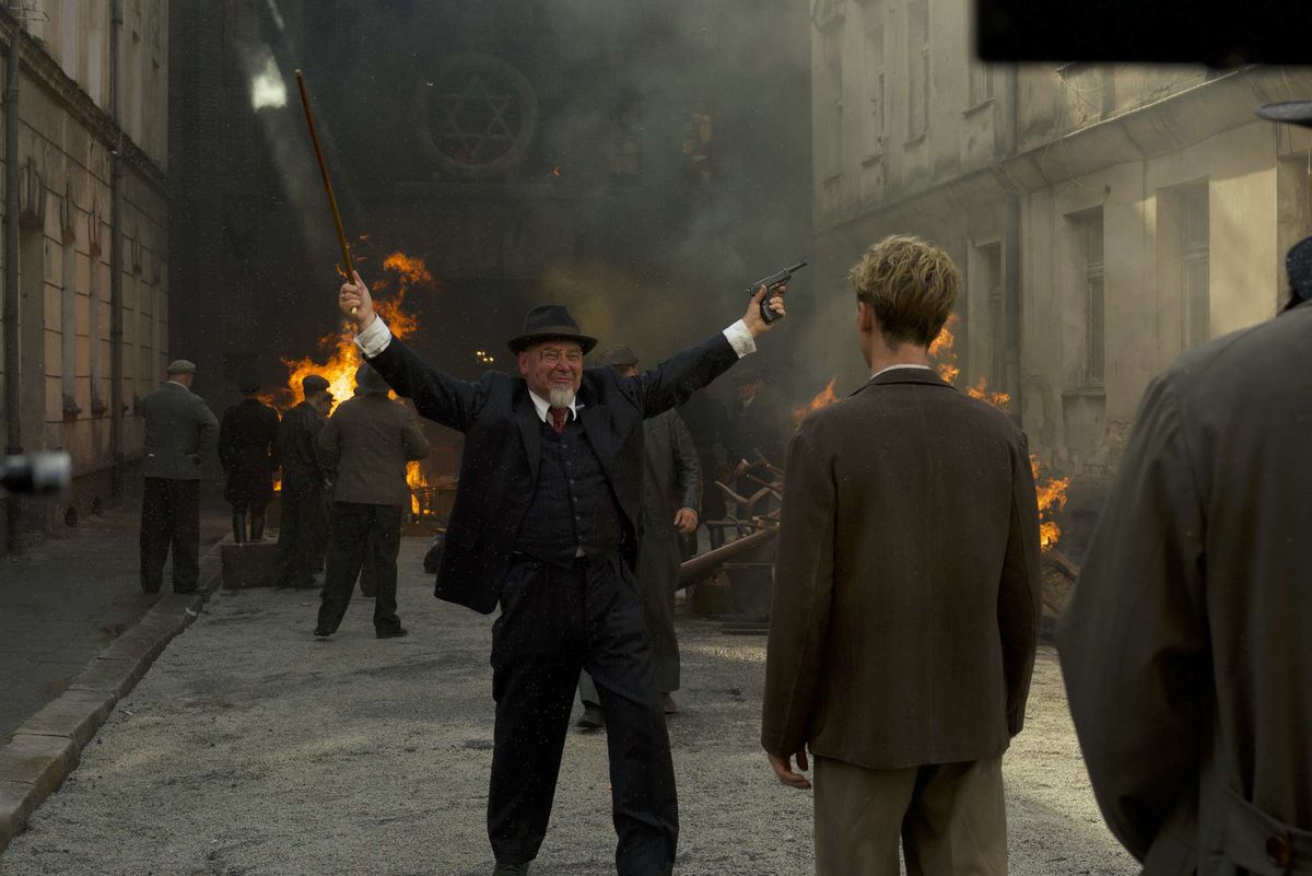 A man in a hat and pointed white beard without a mustache raises his arms triumphantly in front of a burning synagogue.  He is holding a gun