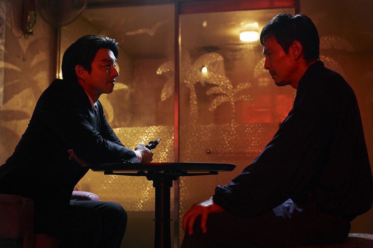 The Recruiter and Gi-hun (played by Gong Yoo and Lee Jung-jae respectively) are sitting at a table, while the Recruiter is holding what looks like a gun pointed away from the table