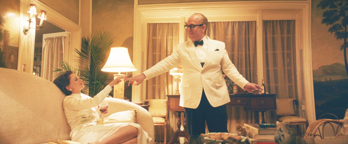 Capote, in a white tuxedo jacket and bow tie, stands above Babe Paley, lounging on a couch in a white dress as he hands her a cigarette in FX's Feud: Capote vs.  The Swans.