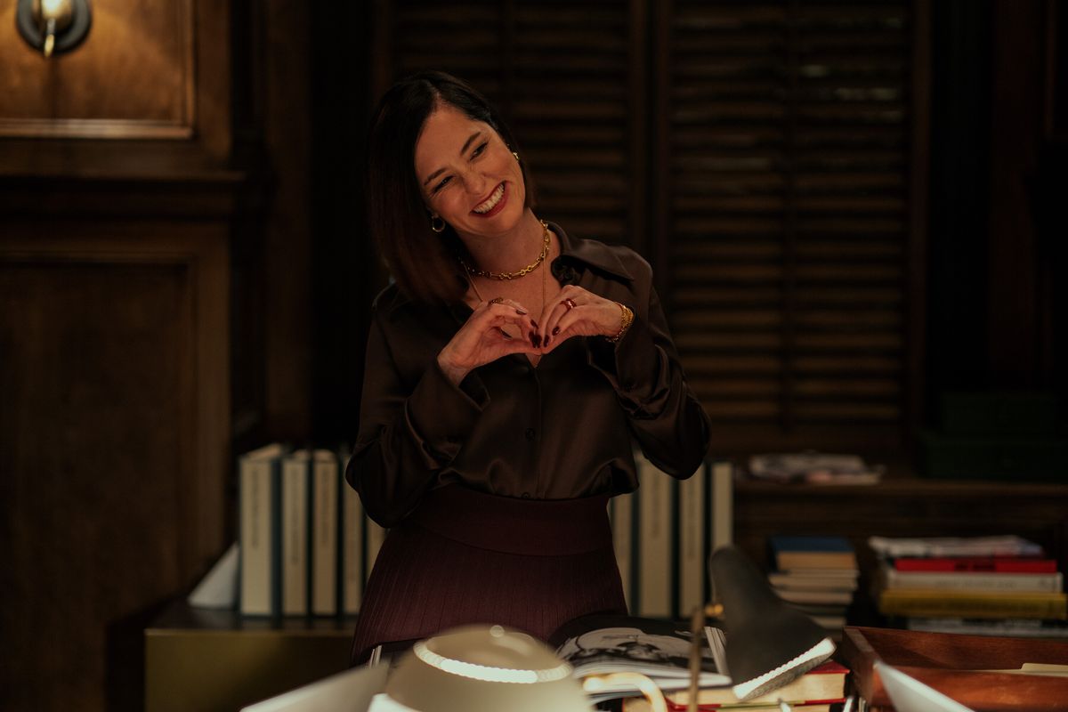 Parker Posey smiles and makes a love heart shape with her hands in a dark study room in Mr.  & Mrs.  Smith