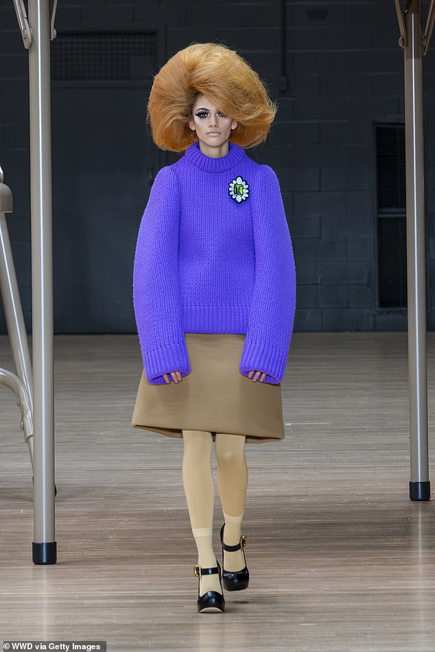 A model with red hair, wearing a purple sweater and a short brown skirt