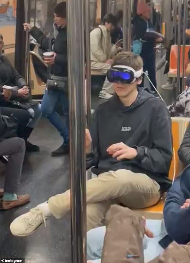 A person wearing brown pants and a gray sweatshirt uses headphones while typing in the air on a subway in New York City