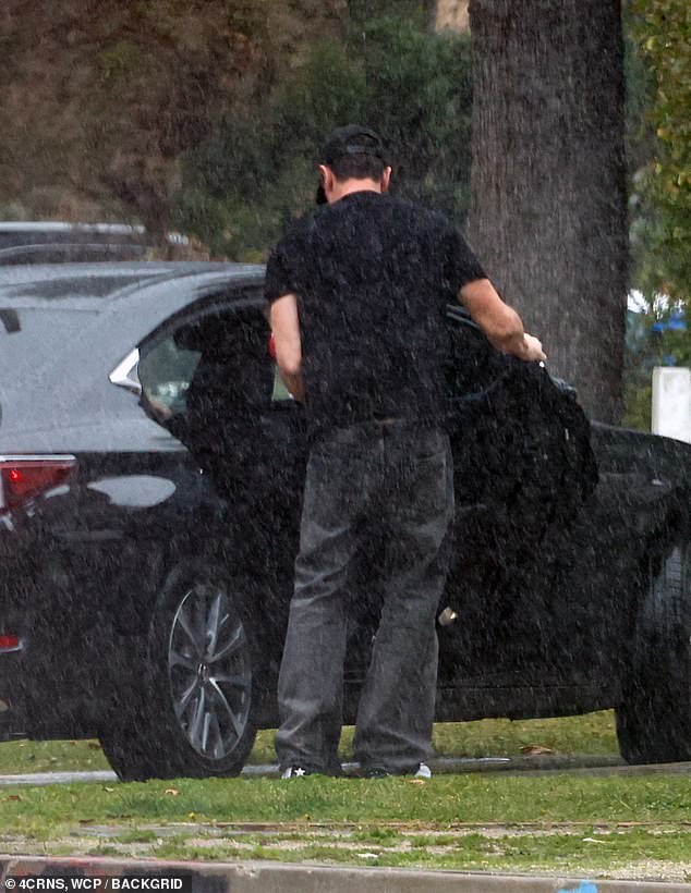 Tom braved the rain in a black t-shirt, oversized dark jeans and a black hat as he carried the flower arrangement