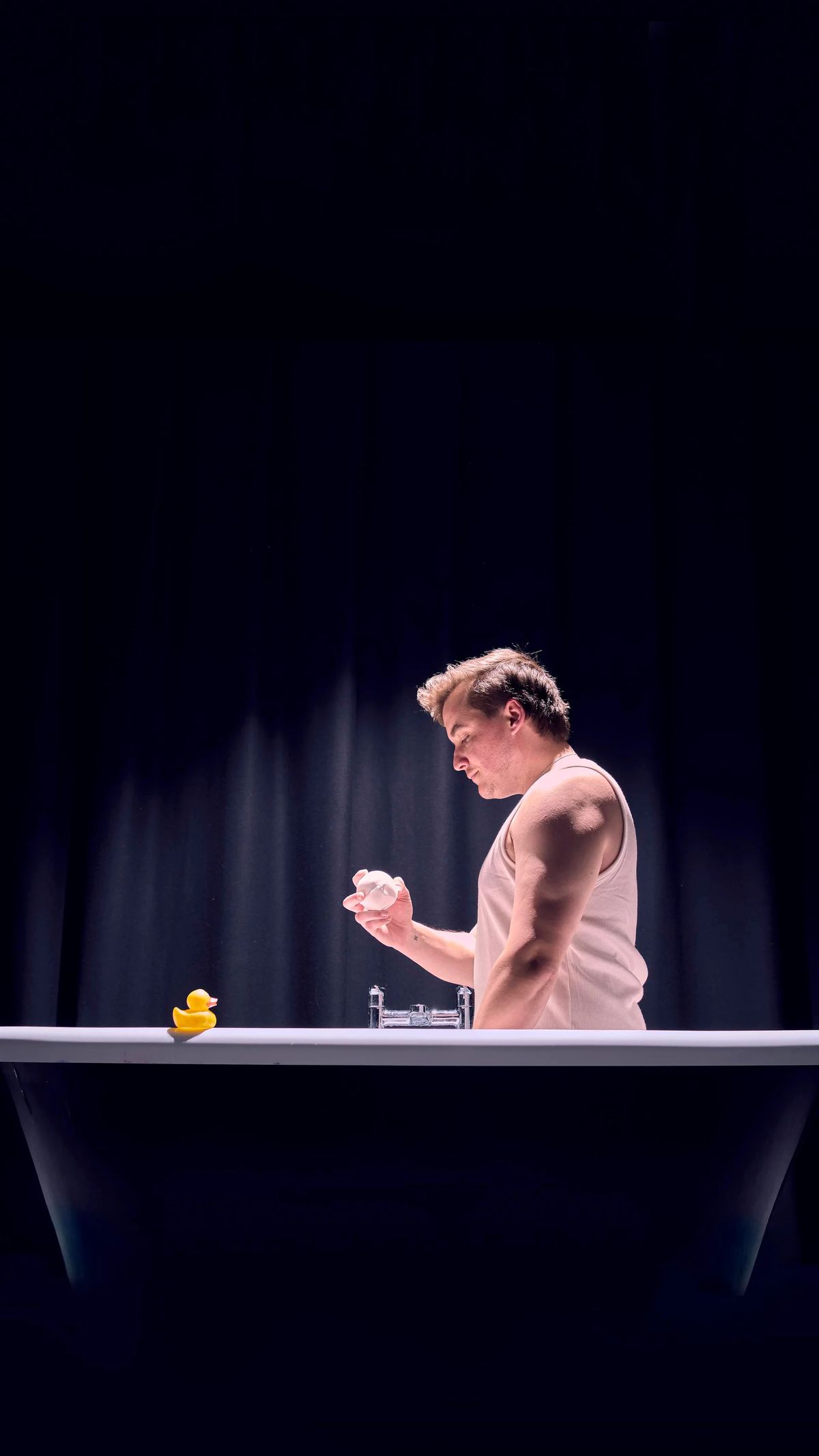 A man stands in a dramatically lit bathtub.  He is wearing a white tank top and holding a white bath bomb and looking at it intensely. 