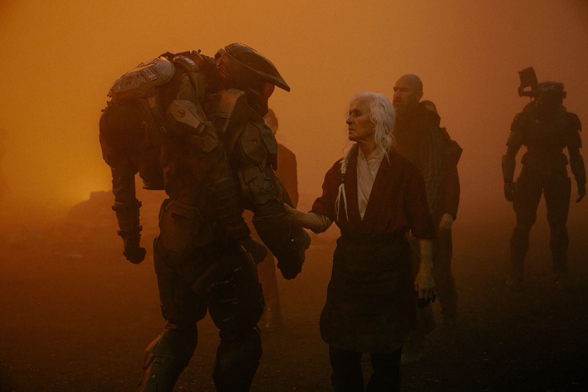 Master Chief carries a wounded Marine over his shoulder as fire and ash cover the sky in Halo Season 2