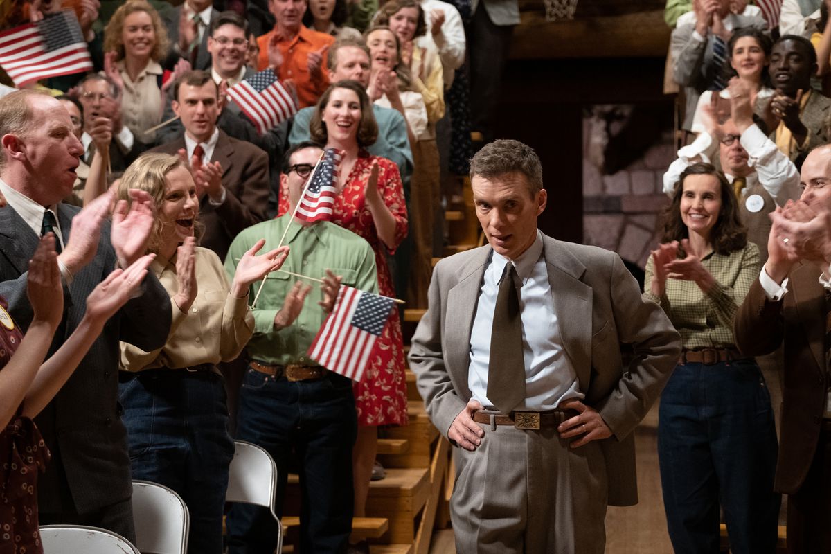 J. Robert Oppenheimer stands with his hands on his waist as he walks between two packed bleachers full of cheering people waving American flags in a scene from the movie Oppenheimer.